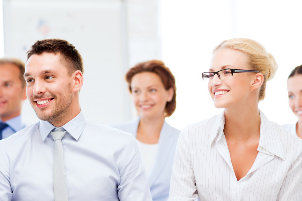 Eine Gruppe professionell gekleideter Menschen, darunter ein Mann und eine Frau im Vordergrund, sitzen lächelnd in einer hellen Büroumgebung - vielleicht nach einem Deutschkurs im Caesar Sprachinstitut.