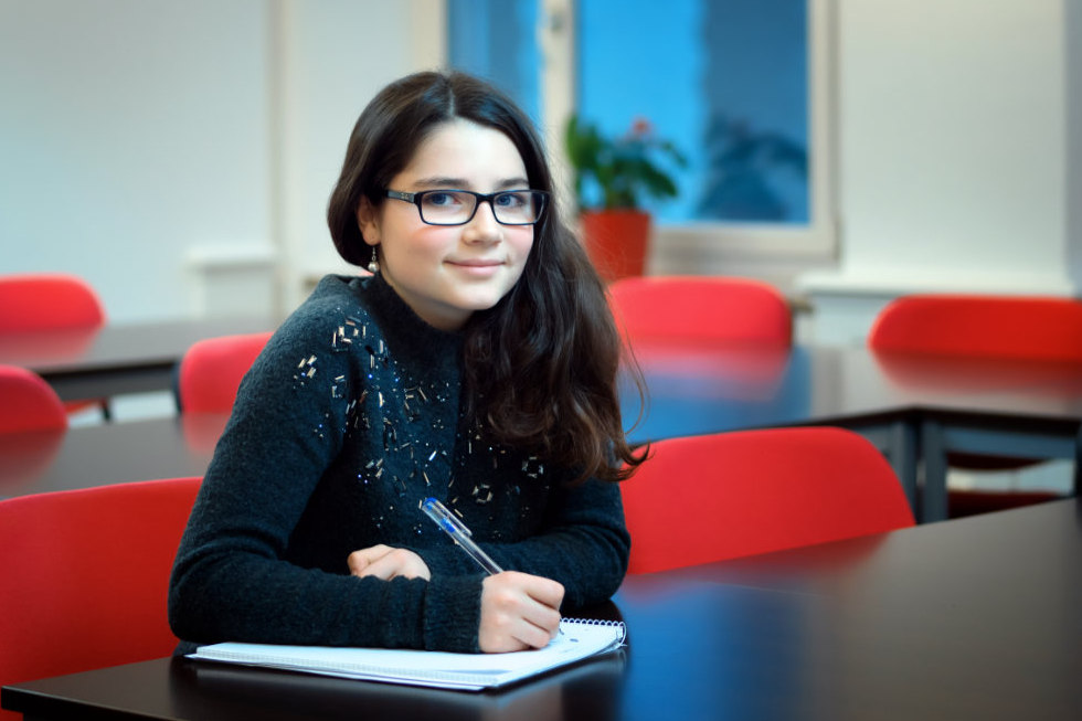 Eine junge Person mit langen dunklen Haaren und Brille sitzt an einem Schreibtisch in einem Klassenzimmer des Caesar Sprachinstituts und schreibt mit einem Stift in ein Notizbuch - vielleicht bereitet sie sich auf eine bevorstehende Sprachprüfung vor. Im Hintergrund sind rote Stühle und ein Fenster zu sehen.
